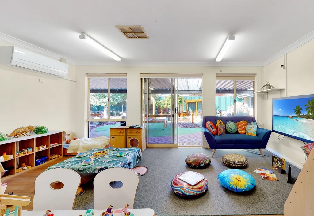 A children's playroom with a couch, TV, table, and various toys. Large windows and a glass door open to a backyard with a patio. The room is lit by ceiling lights.