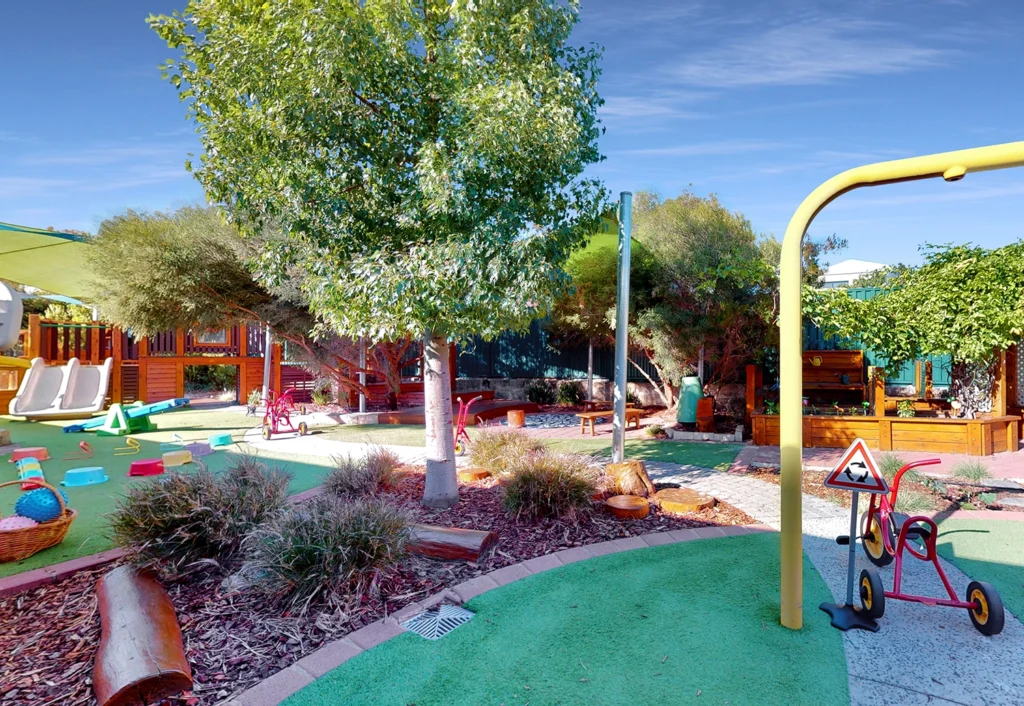 Outdoor playground with green turf, slides, tricycles, and various play equipment surrounded by trees and plants on a sunny day.