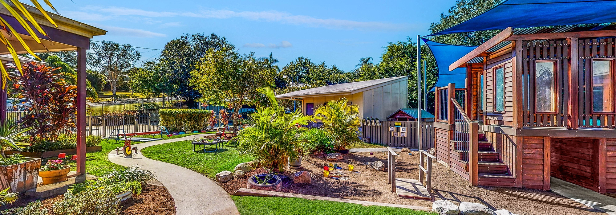 A vibrant playground perfect for early learning, with colorful play structures, green grass, shaded areas, and surrounding trees under a bright blue sky.