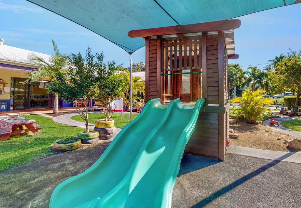 A playground with two turquoise slides, a shaded playhouse, and surrounding greenery including trees and shrubs. In the background, a building with a purple awning is visible.