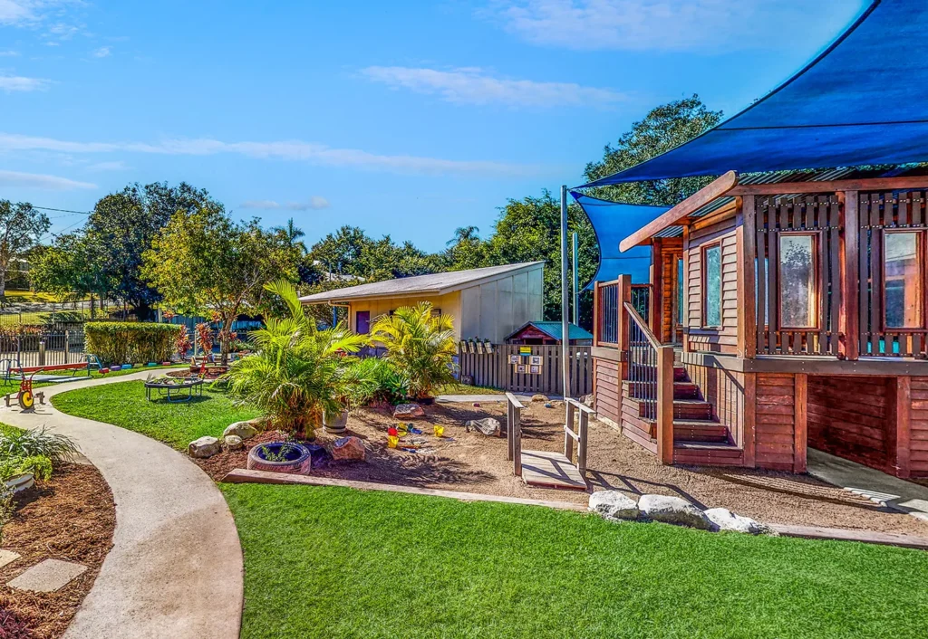 A colorful outdoor playground features a path, lush greenery, a small garden, and a wooden playhouse with blue shade covers against a sunny backdrop.