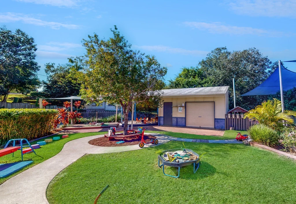 Outdoor playground with green grass, walking paths, a small shed, various play equipment including a tricycle, rockers, and a sandbox. Trees and shade structures are present in the background.