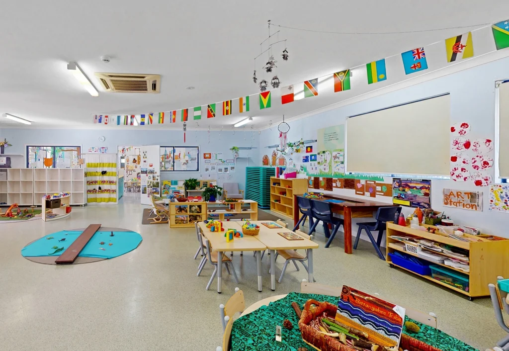 A brightly lit classroom with various learning materials, colorful art on the walls, small tables and chairs, and international flags hanging from the ceiling.