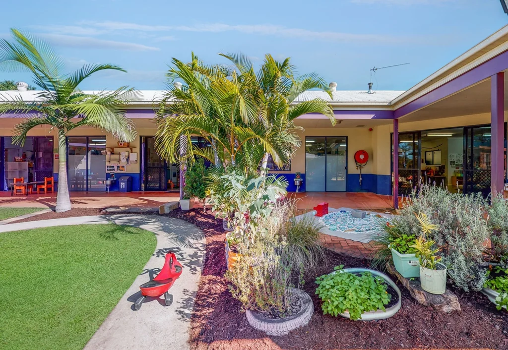 An outdoor courtyard with tropical plants, a small garden, and children's play equipment under an awning with classrooms visible in the background.