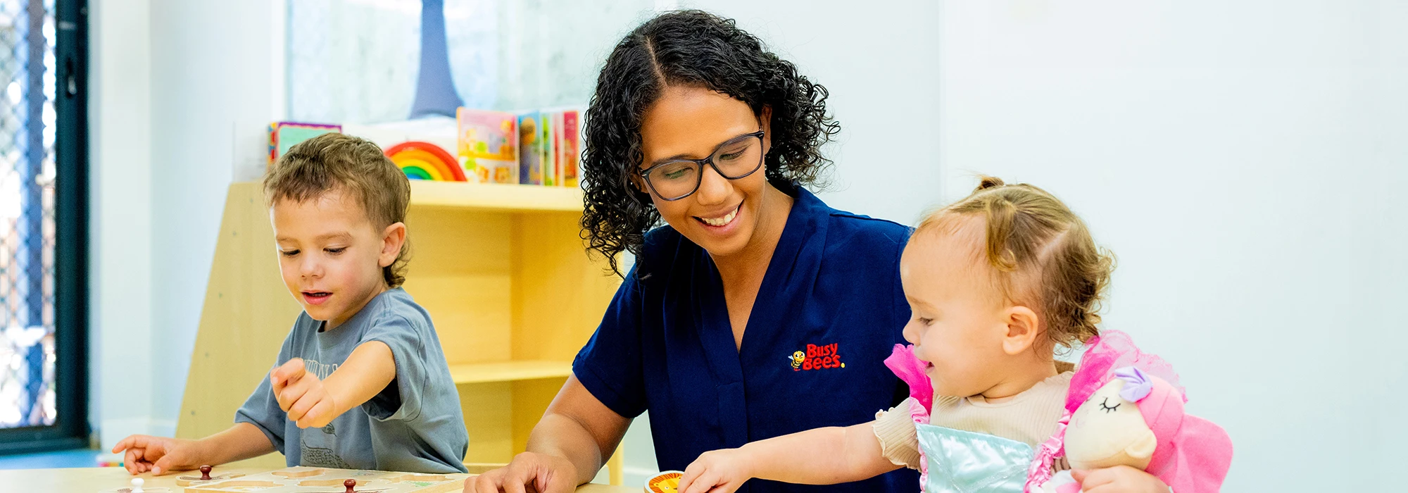 An early childhood educator at Kalgoorlie Boulder day care helps two young children with an activity at a table.