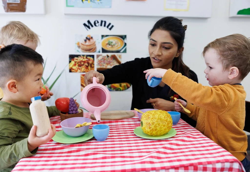 A group of young children and a caregiver engage in tea party play at a table with a red and white checkered tablecloth.