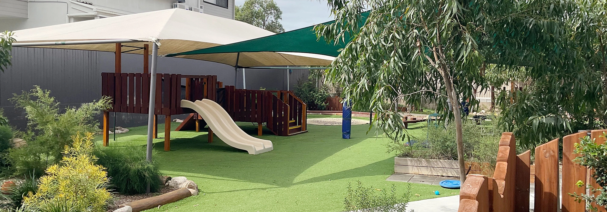 A playground with a slide and climbing equipment under shade structures. The area is surrounded by greenery with artificial turf flooring.