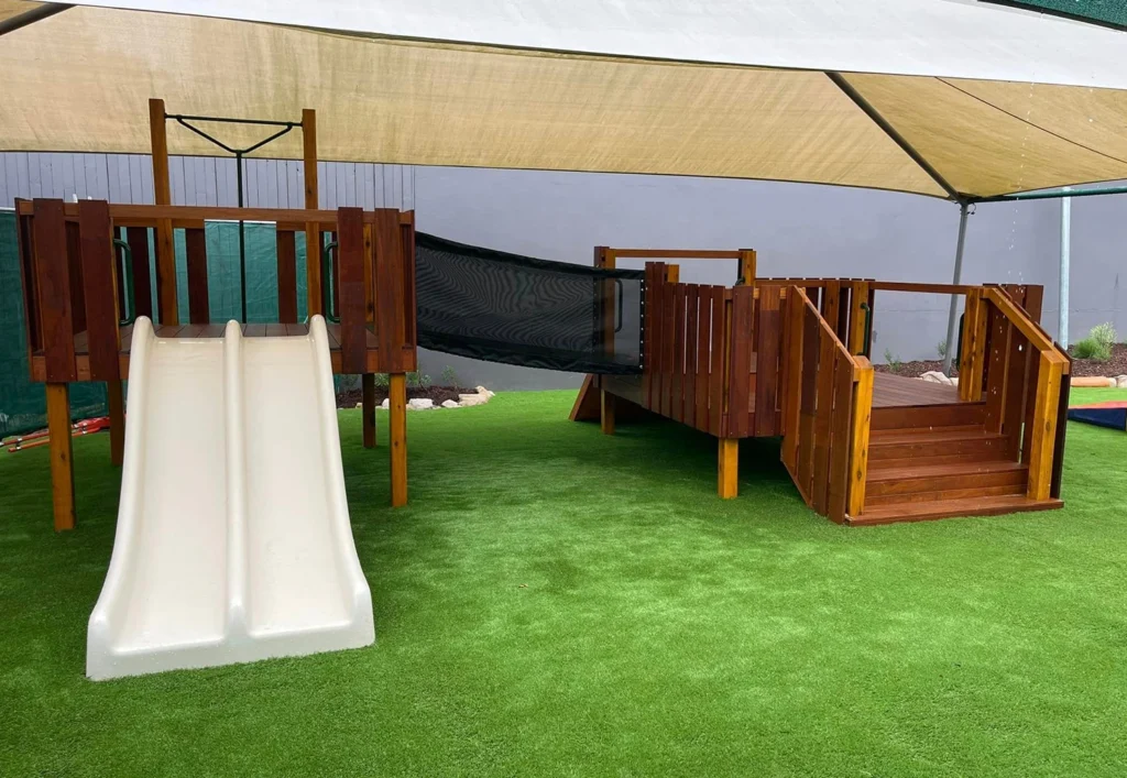 A playground structure with a white slide, wooden platforms, a bridge, and a canopy overhead on a green artificial turf surface.