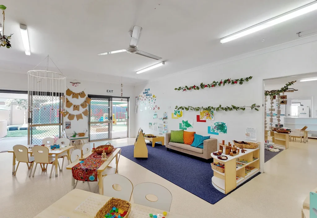 A well-lit daycare center room with child-sized tables and chairs, educational posters on the walls, a sofa on a blue rug, craft supplies, and a play area visible through a glass door.