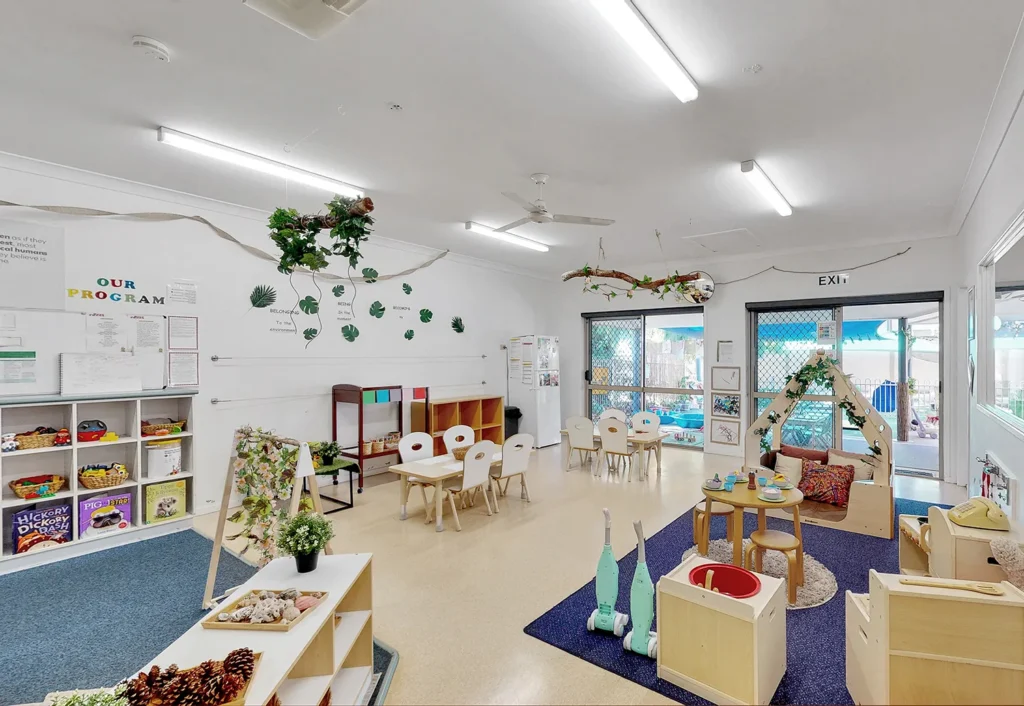 Brightly lit classroom with white walls, children's tables and chairs, bookshelves, and a small play area featuring various toys and decorations. Large windows provide natural light.