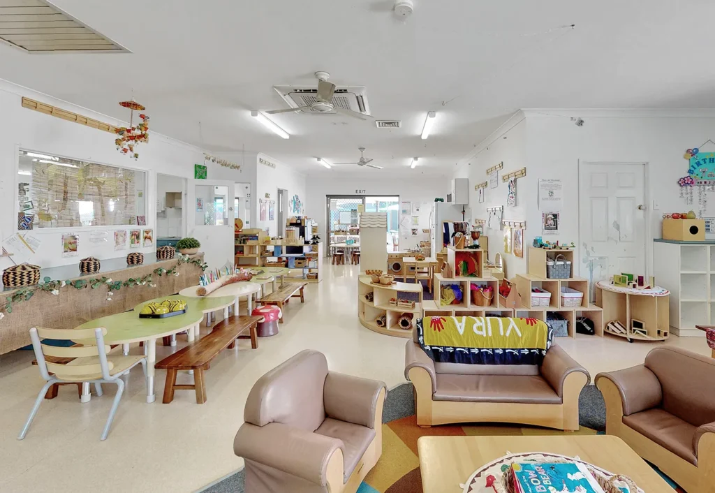 A spacious, well-lit classroom with child-sized furniture, colorful decorations, bookshelves, and various learning stations, arranged neatly throughout the room.