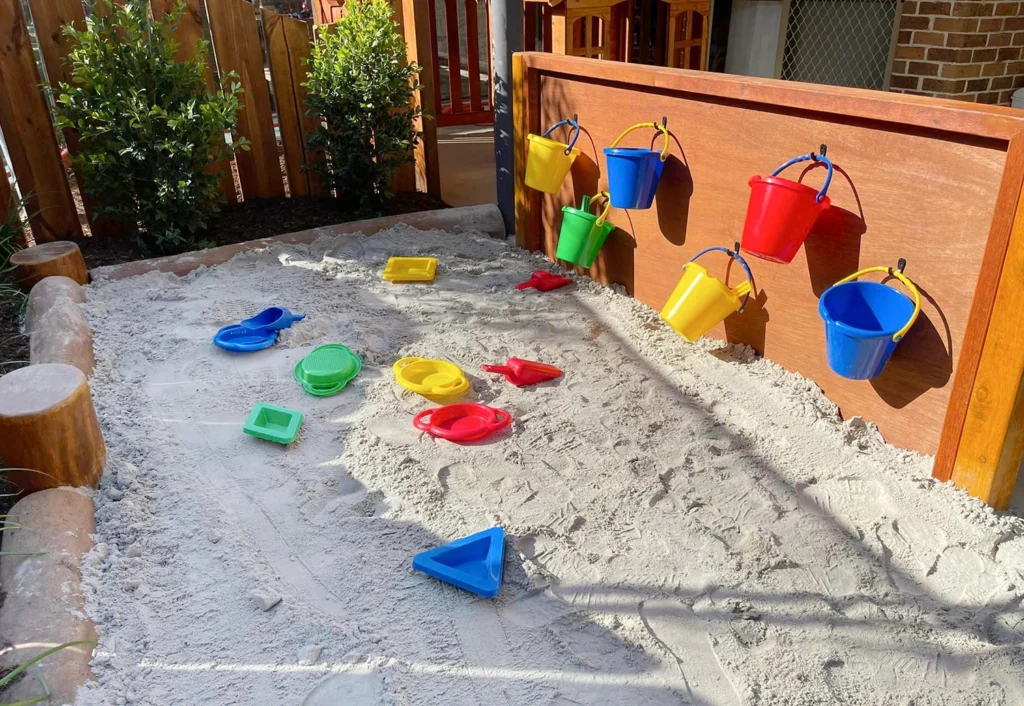 A sandbox with scattered colorful plastic toys including buckets, scoops, and molds. Some buckets are hanging on a wooden board. The area is surrounded by a wooden fence and plants.