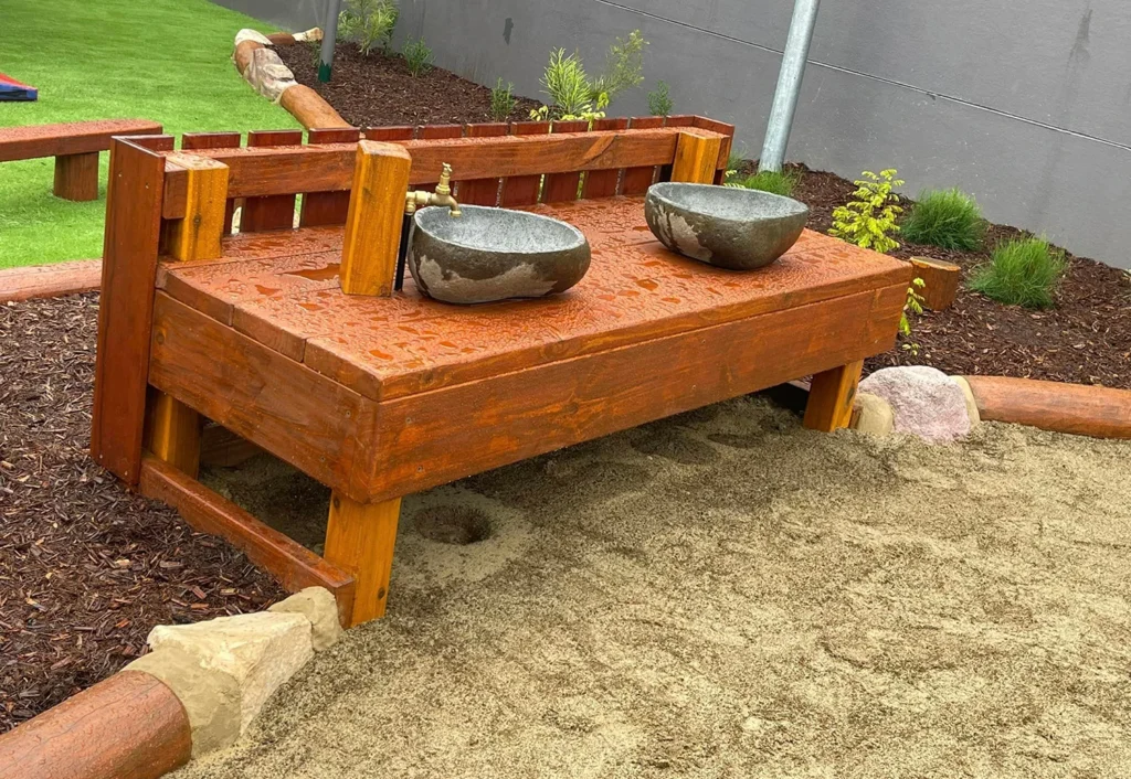 A wooden outdoor playbench with two stone basins and a brass faucet is set in a yard with sand and mulch areas.