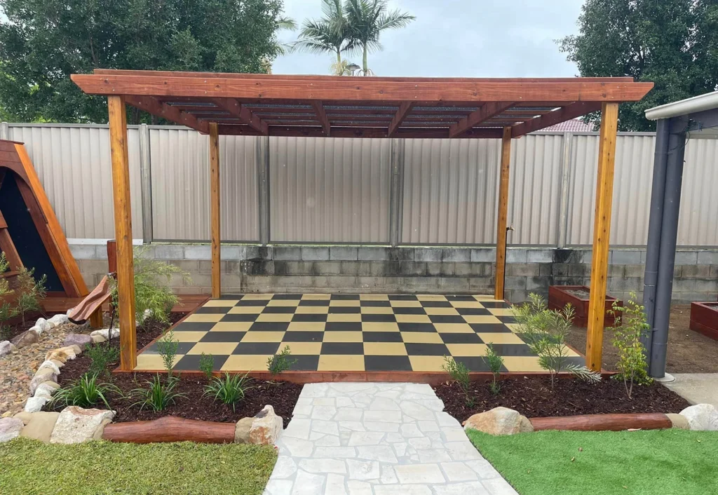 A wooden pergola covers a black-and-tan checkered patio in a backyard. The area is bordered by shrubs and has a pathway leading to it. A tree and a fence are in the background.