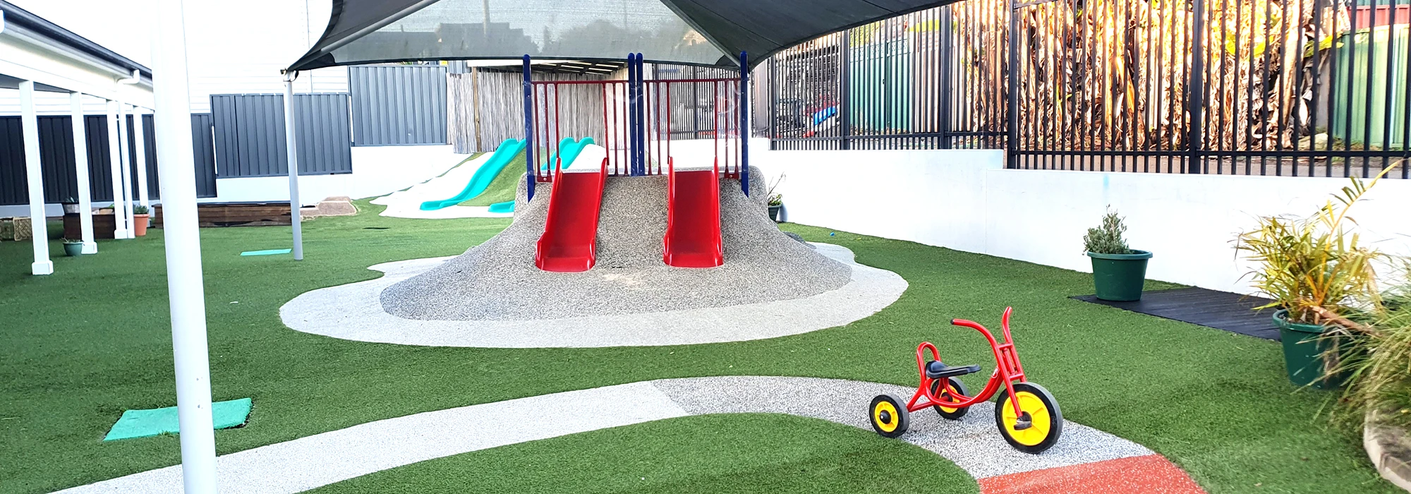 A playground featuring twin red slides, artificial grass, potted plants, a tri-cycle, and a shaded canopy. A climbing structure and a blue slide are in the background.