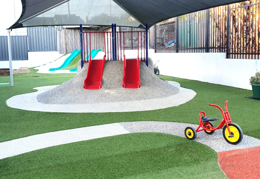 A playground with artificial turf features two red slides on a mound, a shaded area, two green slides in the background, and a red tricycle in the foreground.