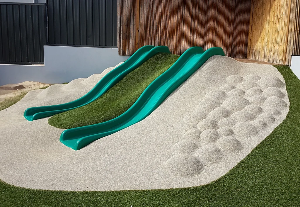 A playground features two green slides side by side on a hill made of artificial turf and sand with bumpy areas for climbing.