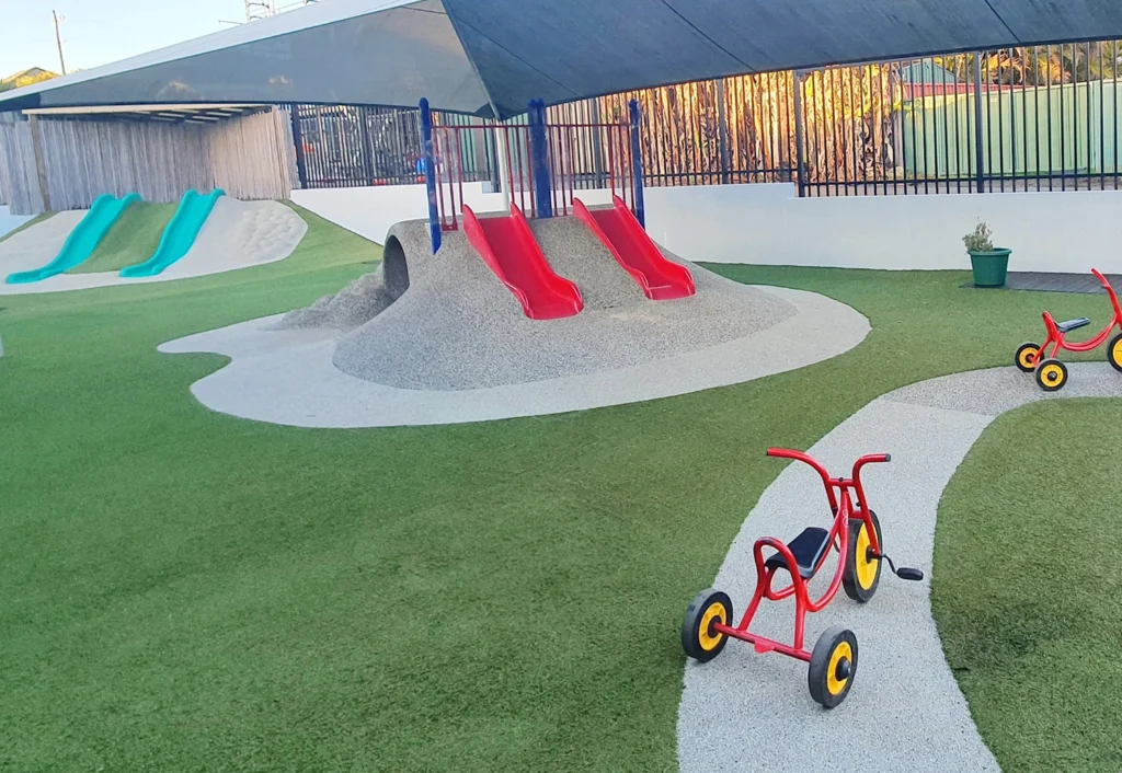 A children's playground featuring red and blue slides, a small hill, green artificial turf, and a couple of red tricycles on a paved path.