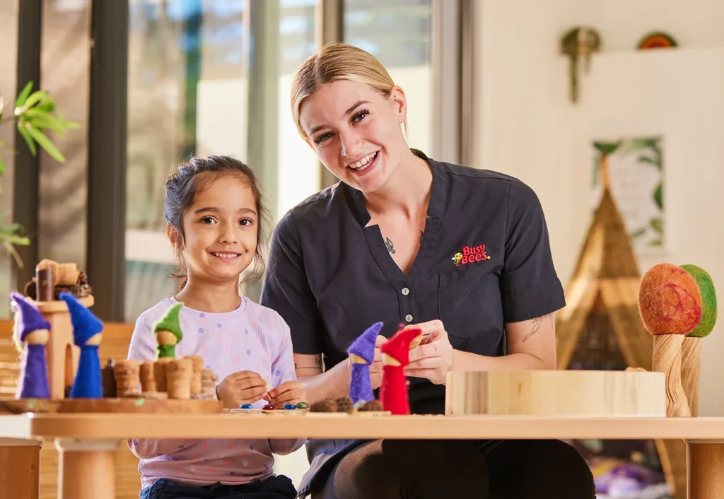 A child and an adult are seated at a table, smiling, and engaged in an arts and crafts activity with colorful materials in a well-lit room.