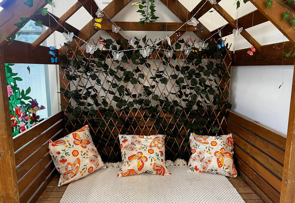 A cozy wooden reading nook with a cushioned seat, three butterfly-themed pillows, and a decorative ivy backdrop adorned with string lights and hanging butterfly ornaments.