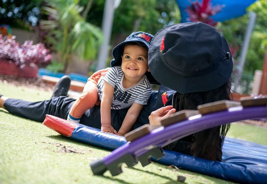 A baby wearing a striped shirt and a black hat with red embroidery smiles while crawling on a mat beside a person lying down, also wearing a similar hat. Outdoor setting with garden background.