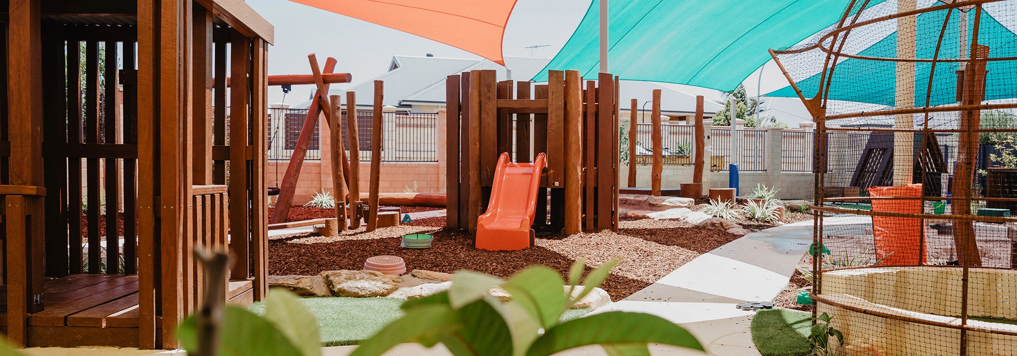 A playground with climbing structures, slides, and shaded areas. It has woodchip ground covering and various play equipment.