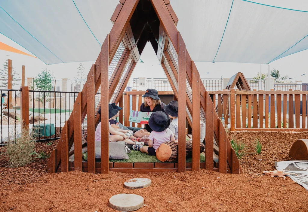 A group of children and a woman are sitting inside a triangular wooden play structure under a shaded area at an outdoor playground. They appear to be engaged in an activity or conversation.