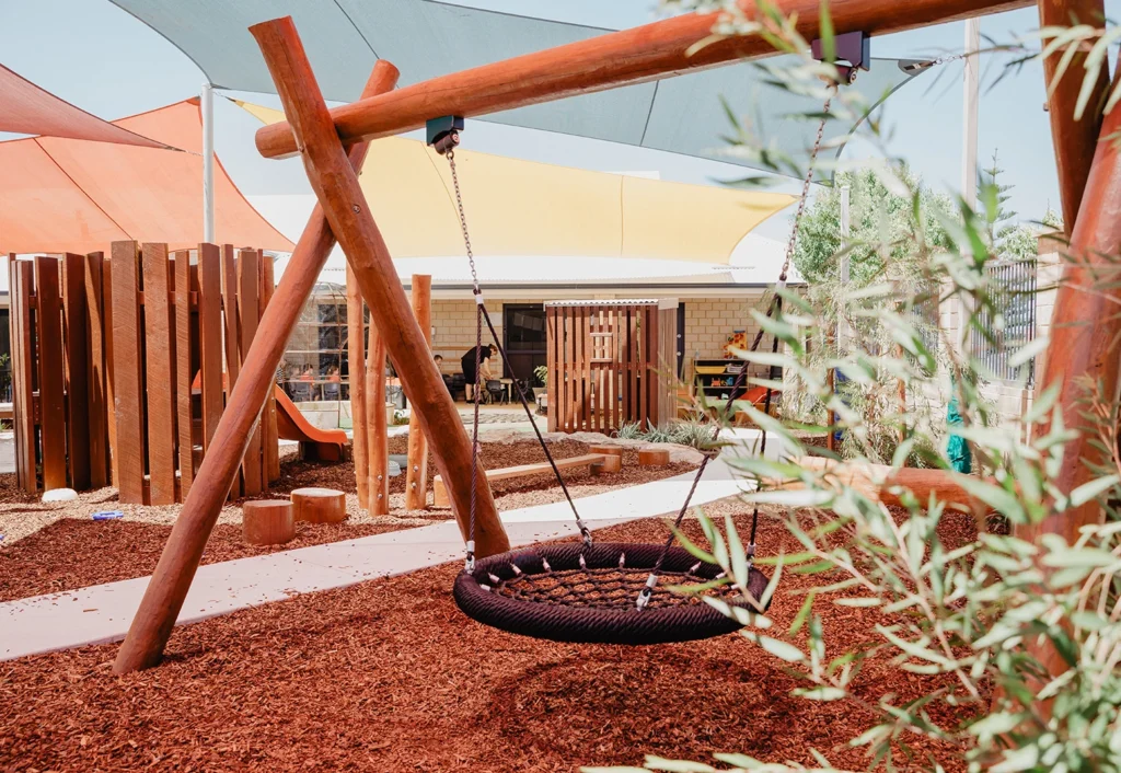 A children's playground with a round rope swing, shade sails, a slide in the background, and wood chip ground cover.