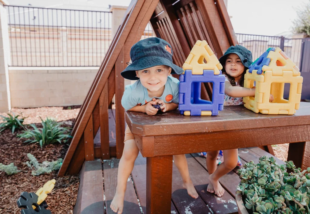 Two children in sun hats play with colorful building blocks on a wooden platform beside an A-frame structure in an outdoor play area.