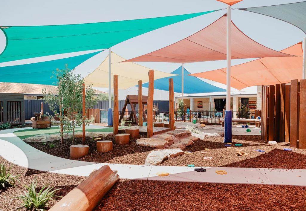 A colorful outdoor playground with various wooden structures, shaded by large, multicolored triangular canopies, surrounded by mulch and small plants.