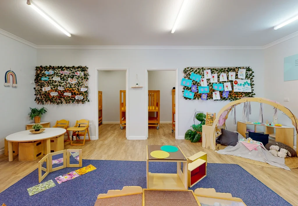 A brightly lit nursery classroom with craft displays on the walls, small wooden tables and chairs, and a play area with toys and books on the floor.