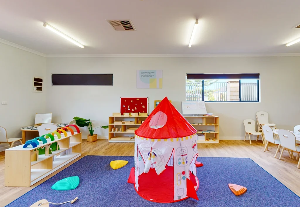 A brightly lit playroom featuring a red and white rocket-shaped tent, colorful toys on shelves, and child-sized chairs arranged along the walls. The floor is covered with a blue rug.