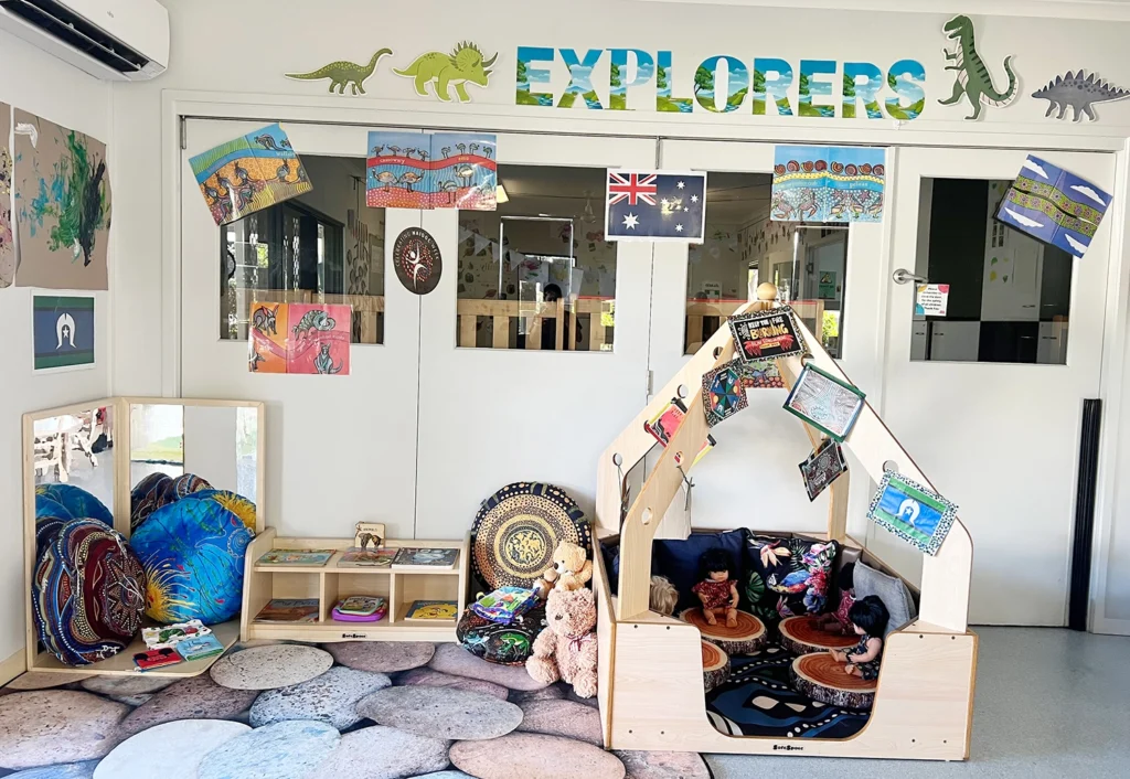 A children's reading corner with a tent, stuffed toys, and books. Decor includes dinosaur cutouts, phrase "EXPLORERS," and various artworks on the wall. Multi-colored cushions are on the floor.