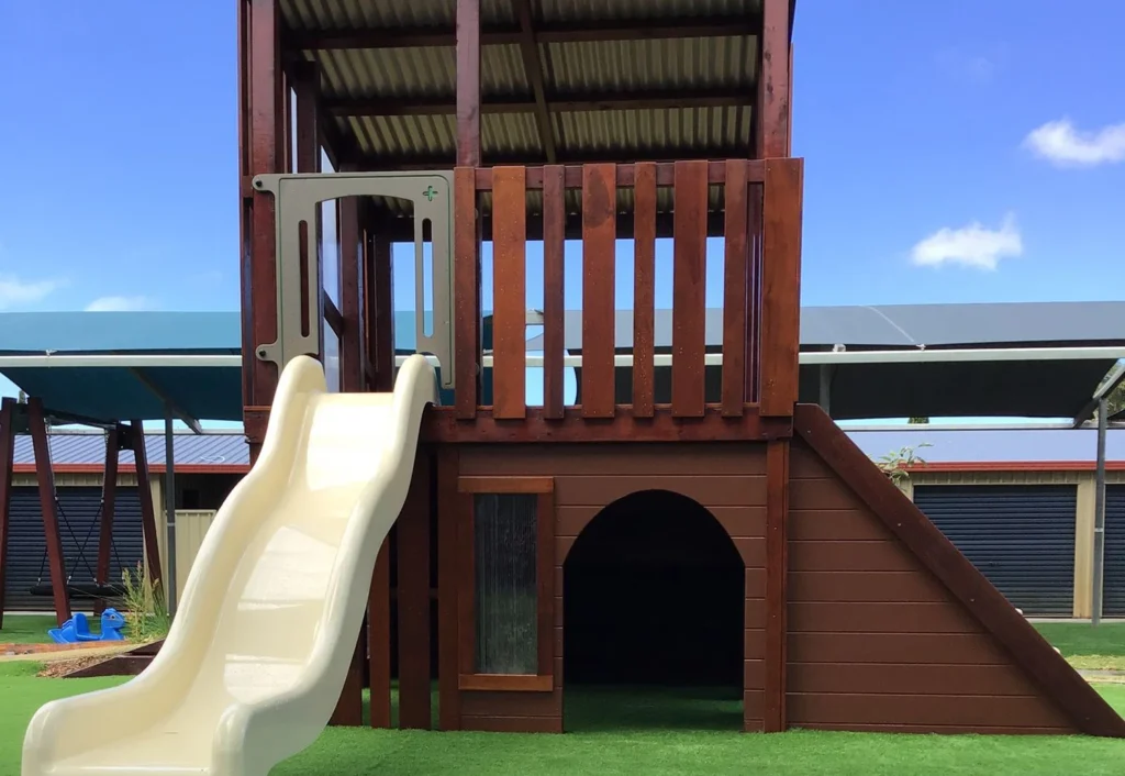 A wooden playground structure with a beige slide, stairs, and a small tunnel entrance underneath, set on artificial grass with a clear blue sky in the background.