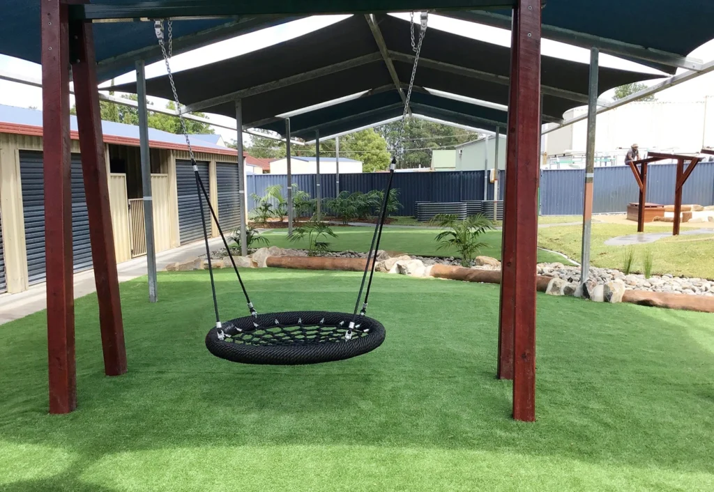 A black circular swing hangs from a wooden structure on artificial grass under a shaded area, with buildings and plants in the background.