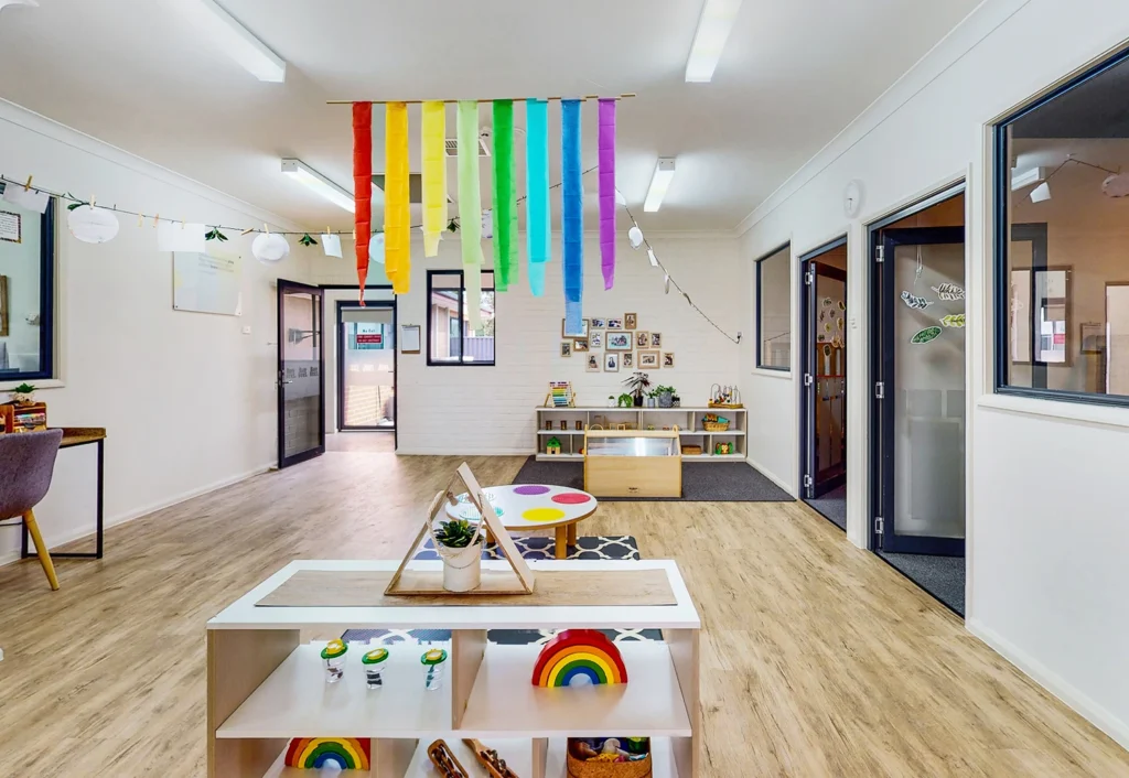 A classroom with wooden flooring and colorful rainbow decorations hanging from the ceiling. Shelves with toys and educational materials are organized throughout the room, with framed artwork on the wall.