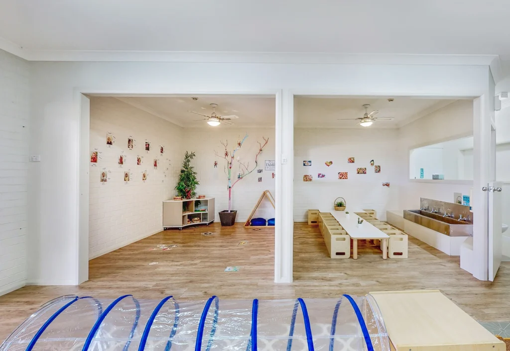 A children's playroom with wooden floors, tables and chairs, a wall with various decorations, a small tree, and a few toys and play structures throughout the room.