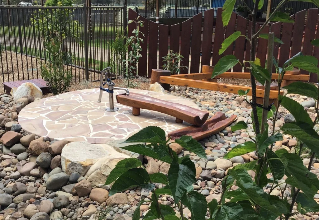 A landscaped garden features a stone-paved area with a wooden water pump and a wooden seesaw-like structure among rocks and plants, enclosed by fencing.