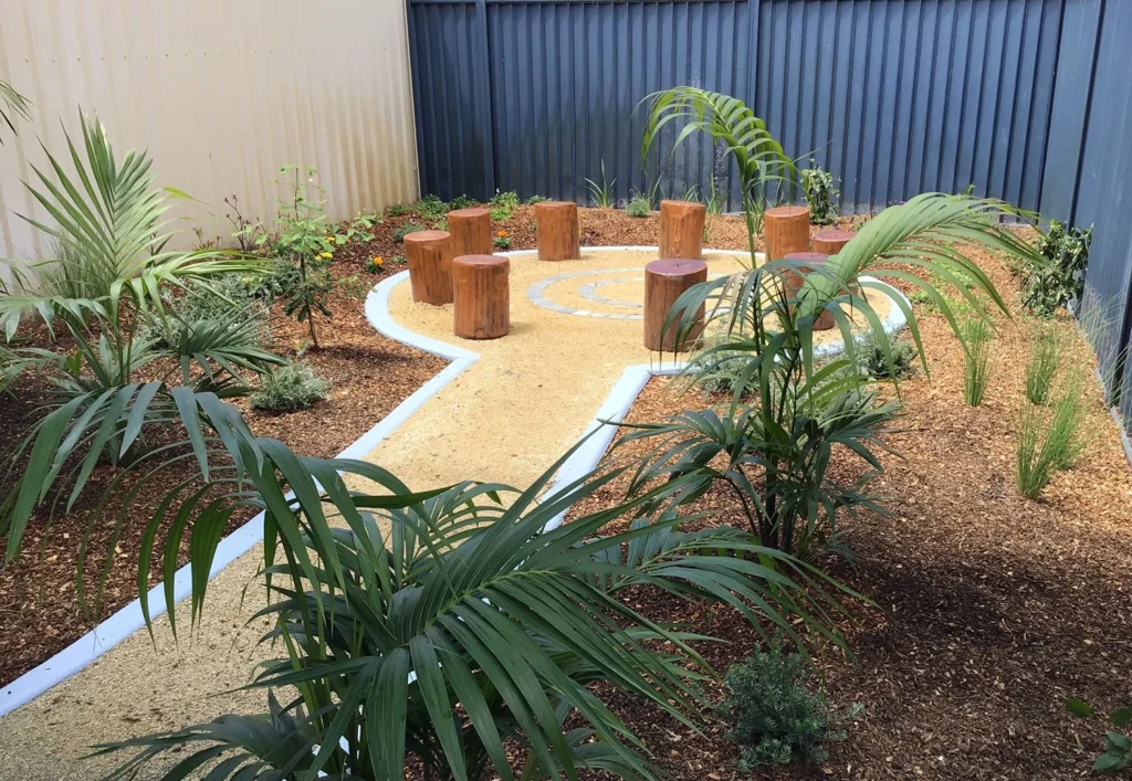 A small, landscaped garden features a gravel pathway, assorted plants, and several wooden stump seats arranged in a semi-circle. The area is enclosed by metal fencing.