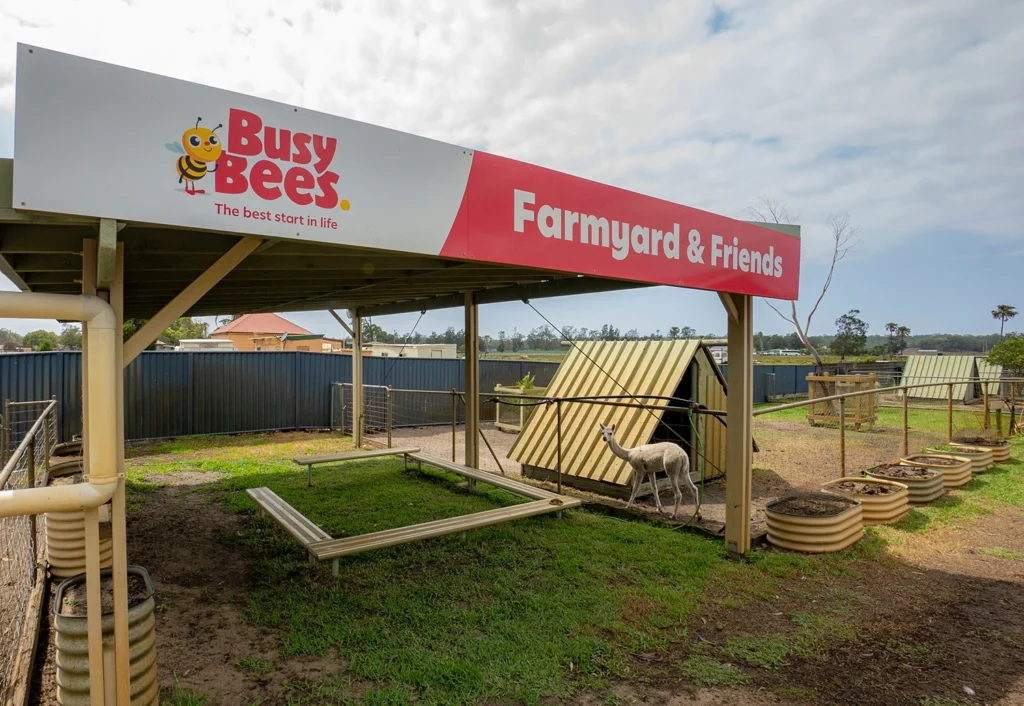 A farmyard area with animal enclosures under a roofed shelter. The shelter has a sign that reads "Busy Bees Farmyard & Friends." A few animals, including a llama, are visible in the enclosure.