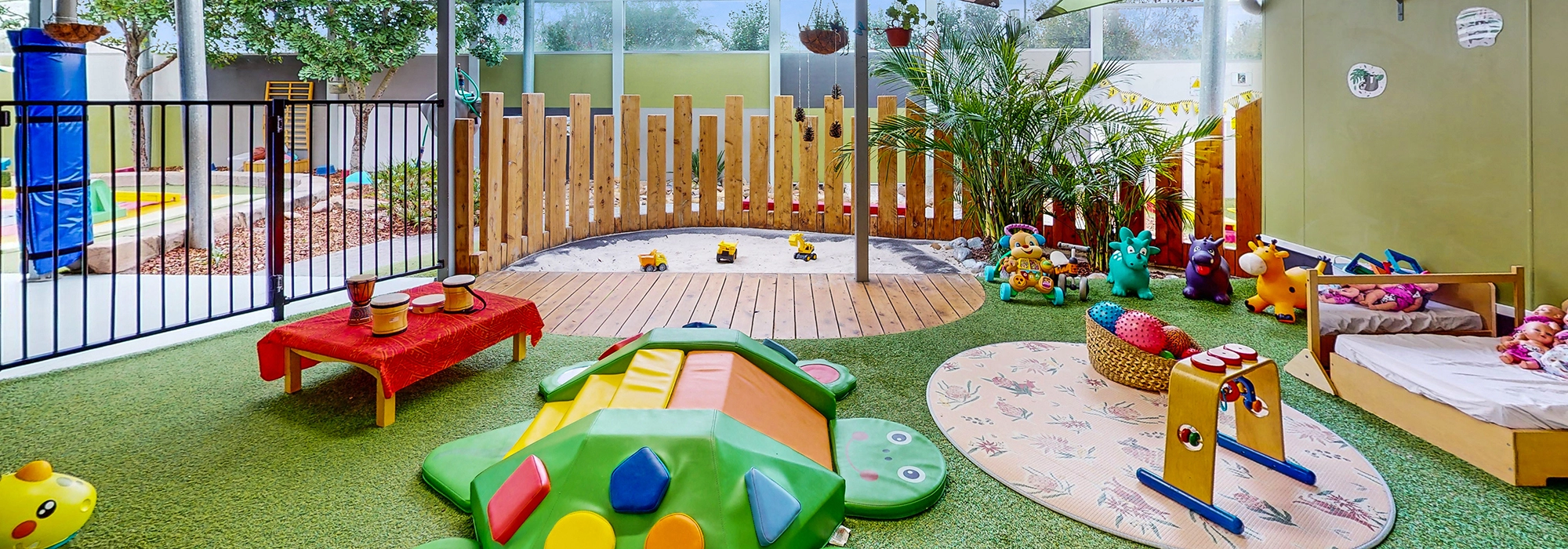 A brightly colored daycare play area with various toys, a small wooden fence, and artificial grass. The room has large windows and potted plants, creating a cheerful early learning environment for children.