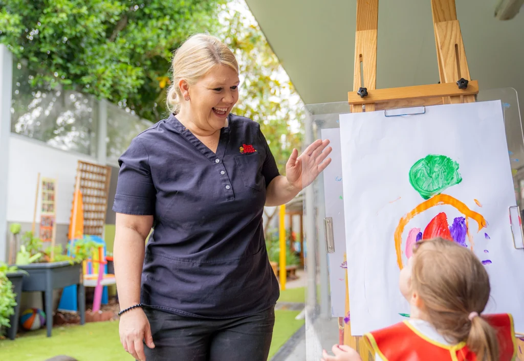 A woman in a dark top smiles and high-fives a young child in a red apron, who is proudly showing a colorful painting on an easel outdoors at the preschool. Trees and playground equipment are visible in the background.