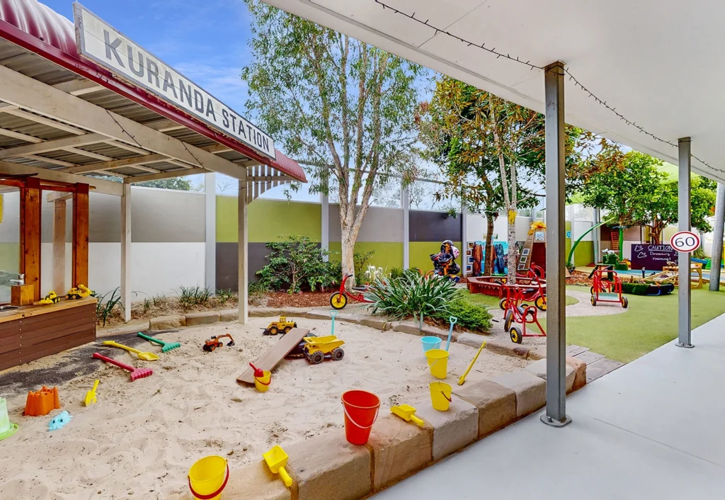 The outdoor children's play area, perfect for a kindergarten or preschool setting, features a sandbox with toys, a small play structure resembling a train station, and various playground equipment under a canopy. Trees and a fence provide a safe and charming backdrop.