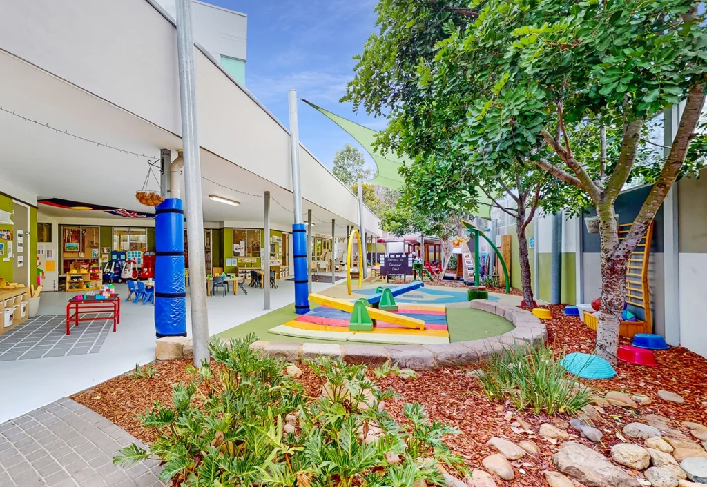 Outdoor area of a preschool features playground equipment, a large shade tree, and various plants. The building has floor-to-ceiling windows and colorful decorations, making it an inviting space for children.