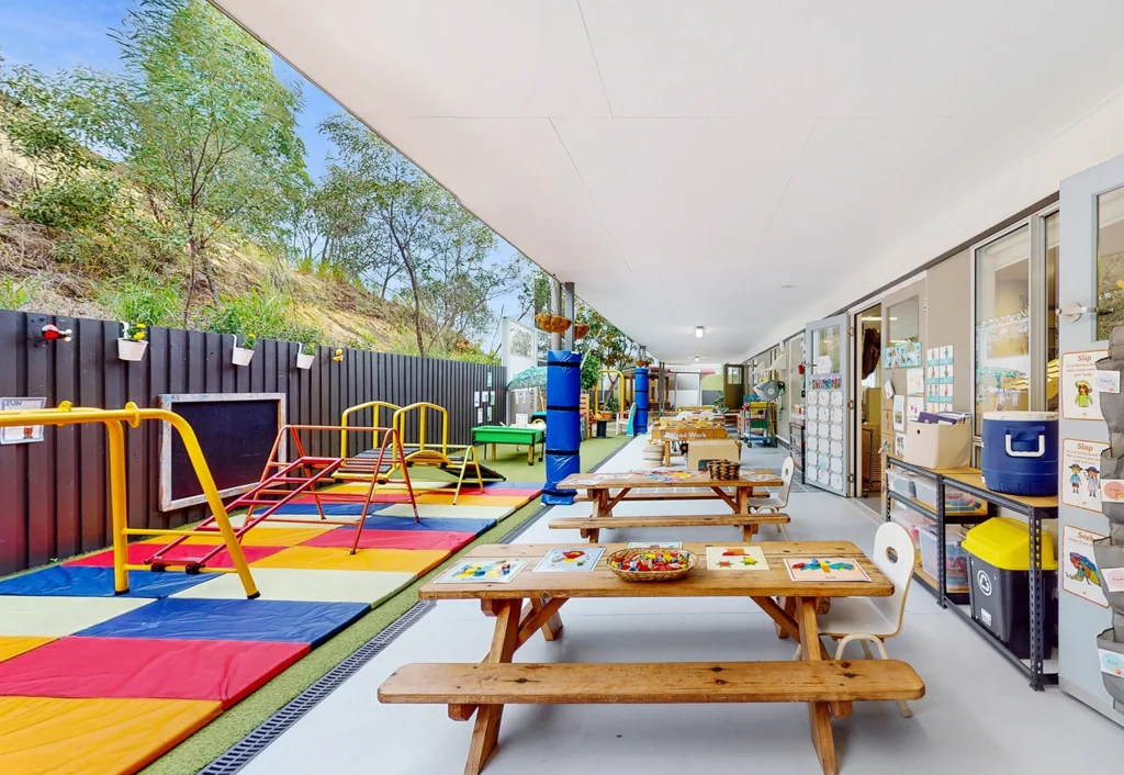 Outdoor early learning area with colorful climbing equipment, padded floor mats, and wooden tables set with craft supplies on a covered patio. Trees and greenery are visible in the background.