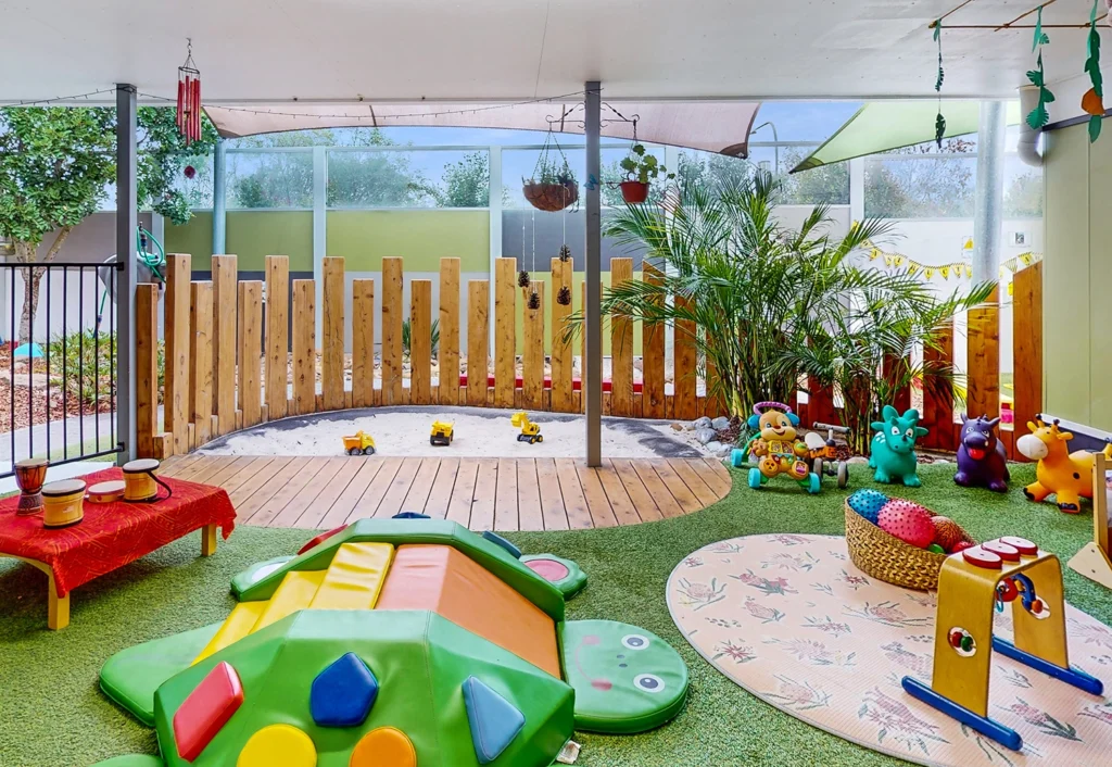 A brightly colored outdoor playground at the preschool features a slide, various toys, and a sandbox surrounded by a wooden fence. Green artificial grass covers the ground, and plants are placed throughout the area, creating an ideal environment for early learning and childcare activities.