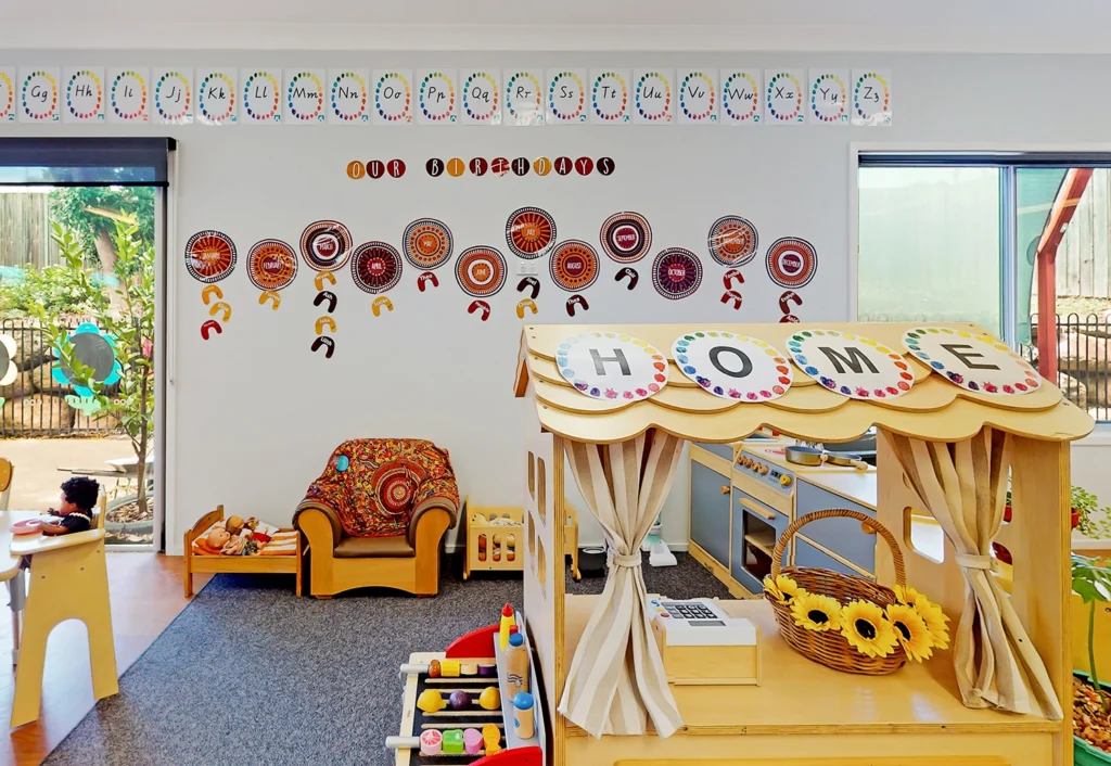 A colorful kindergarten classroom designed for early learning, featuring a play kitchen and shop, wall decorations with a the alphabet and circular patterns, and the word "HOME" displayed on a wooden playhouse structure—perfect for any day care or childcare environment.