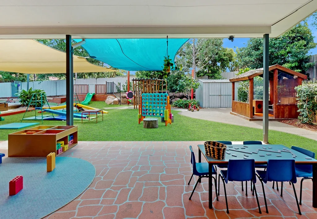 Outdoor playground with various play structures, shaded areas, and a small table with blue chairs. Trees and a fence are in the background, creating an ideal day care environment.