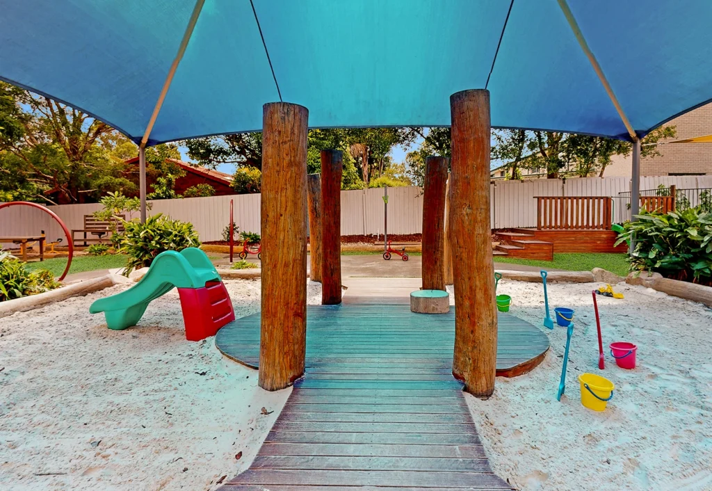 A children's playground, ideal for preschool and kindergarten kids, features a slide, wooden structures, and various sand toys beneath a large blue canopy.