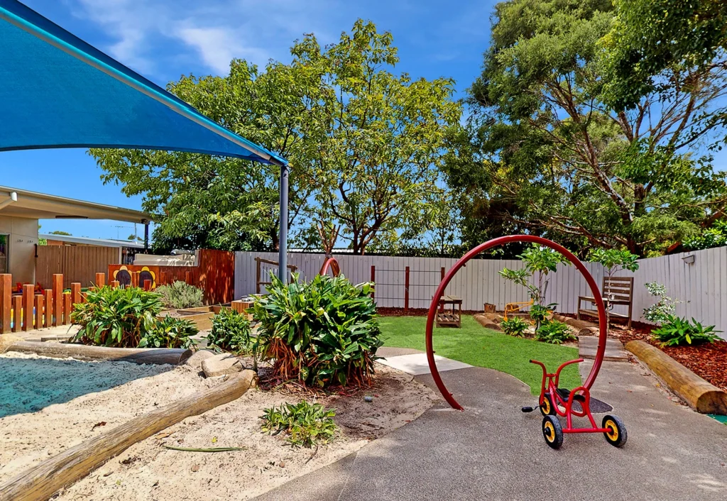 The outdoor playground area at Underwood Kindergarten features a sandpit, a tricycle on a bike track, and a red archway with water misters. The space is surrounded by a white fence and trees, providing a safe and fun environment for young children.